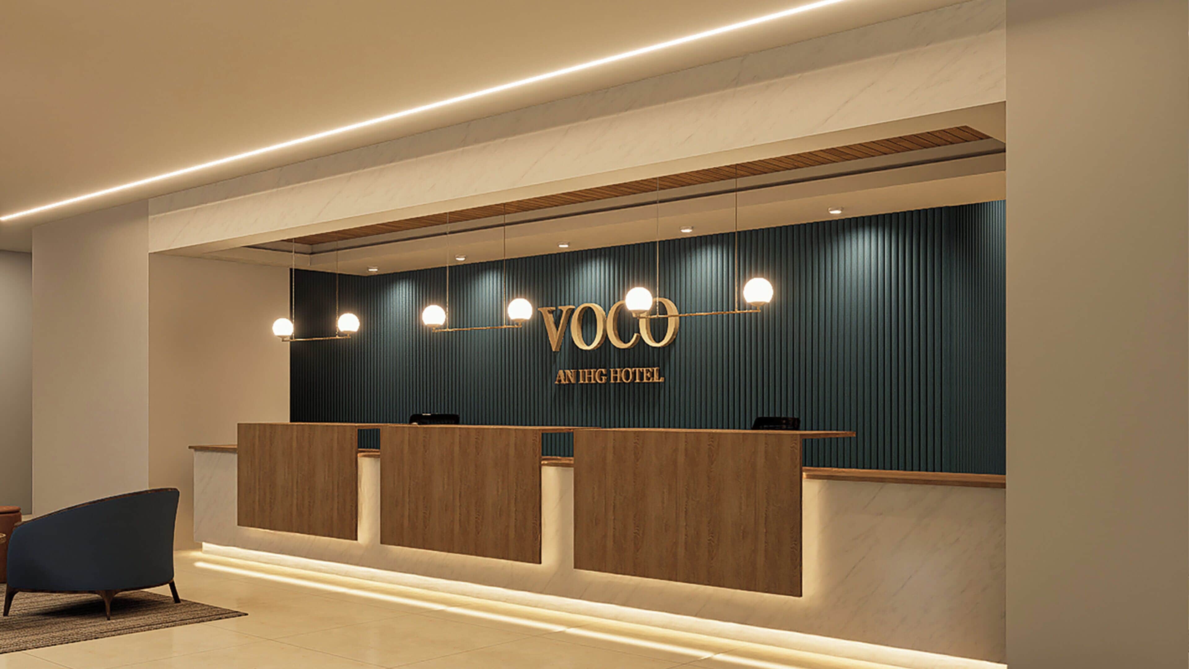 A reception desk with a dark blue wall, wooden panels, and a sign reading 'Voco An IHG Hotel'.