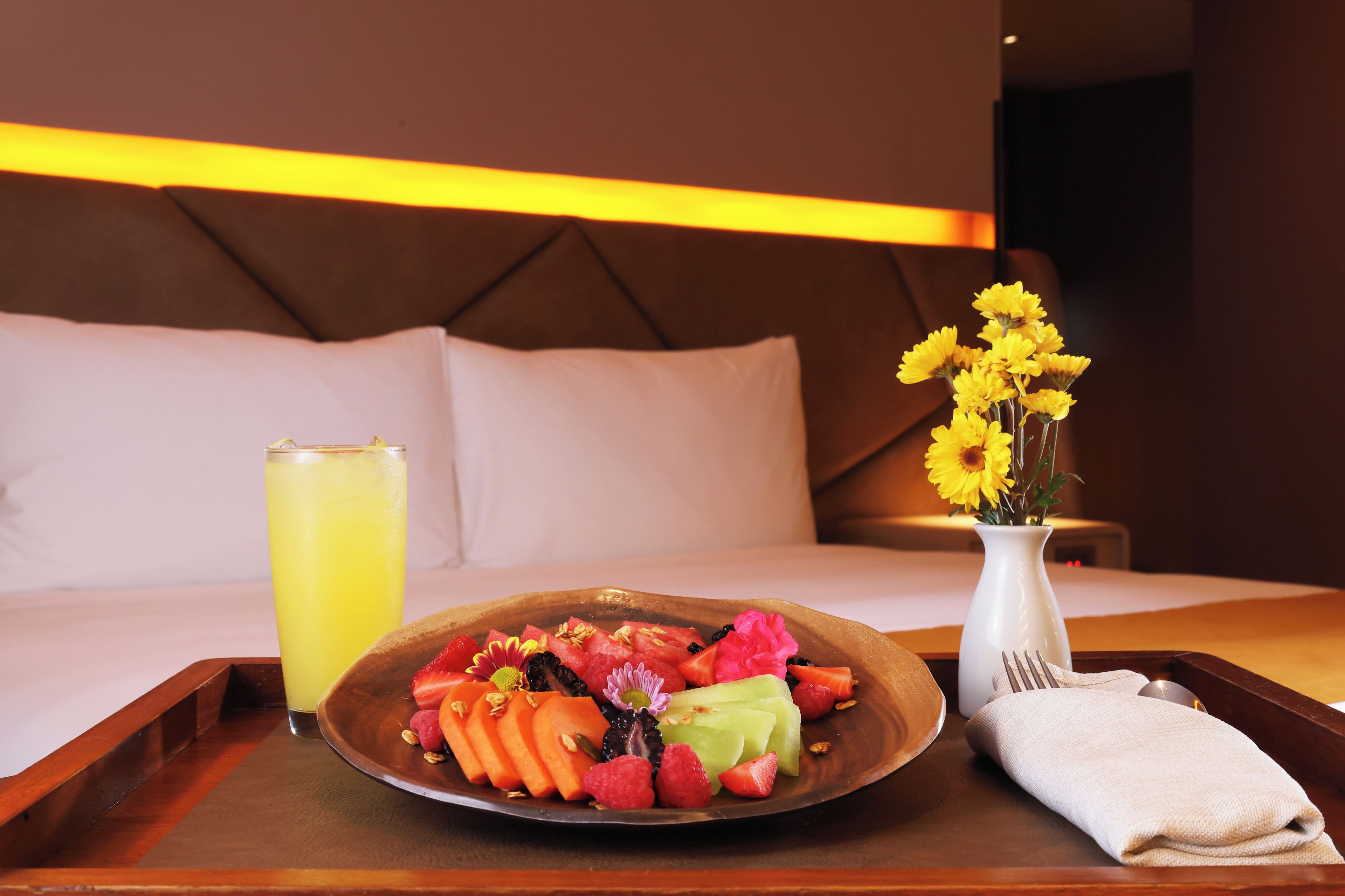 Una bandeja de desayuno bien ordenada con una fuente de fruta, un vaso de bebida amarilla y un jarrón con flores amarillas sobre una mesa de madera en una habitación de hotel.