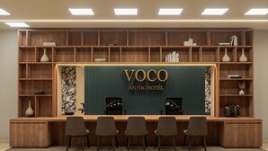 Un mostrador de recepción con un cartel que dice 'VOCO AN IHG HOTEL', estanterías de madera con libros y objetos decorativos, y tres sillas al frente.