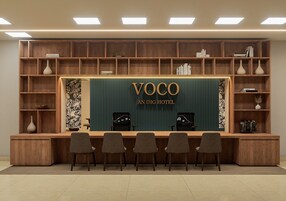 Un mostrador de recepción con un cartel que dice 'VOCO AN IHG HOTEL', estanterías de madera con libros y objetos decorativos, y tres sillas al frente.