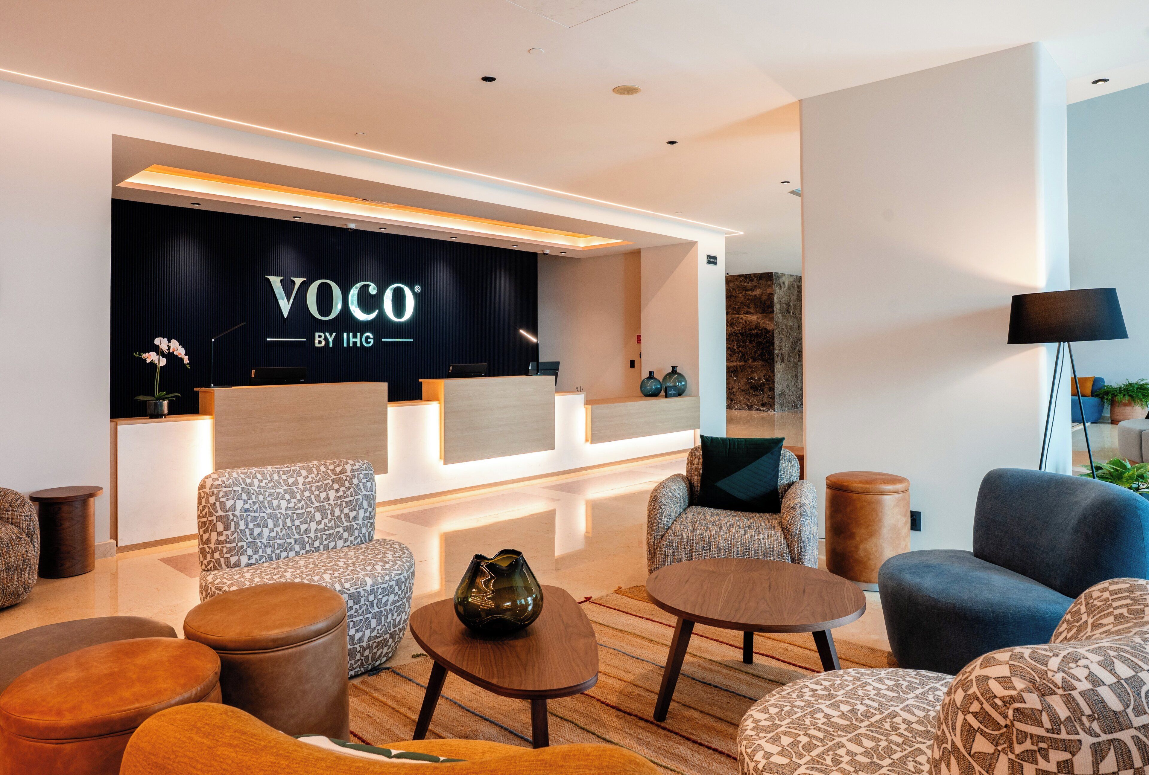 Un hall d’entrée moderne avec des sièges, un comptoir d’accueil et un mur sur lequel est inscrit le texte « Voco by IHG ».
