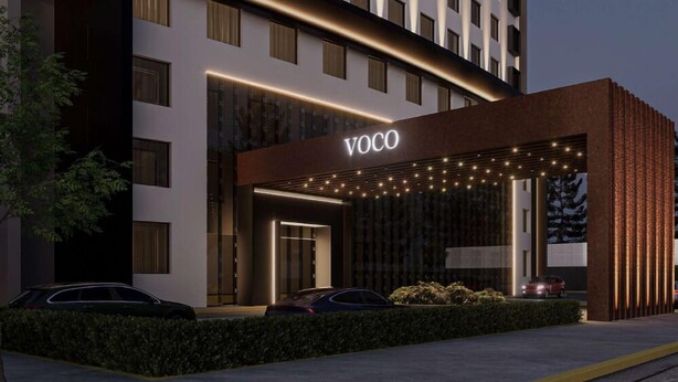 Un hotel moderno con un llamativo letrero 'VOCO', un acceso cubierto y un área de estacionamiento con automóviles.