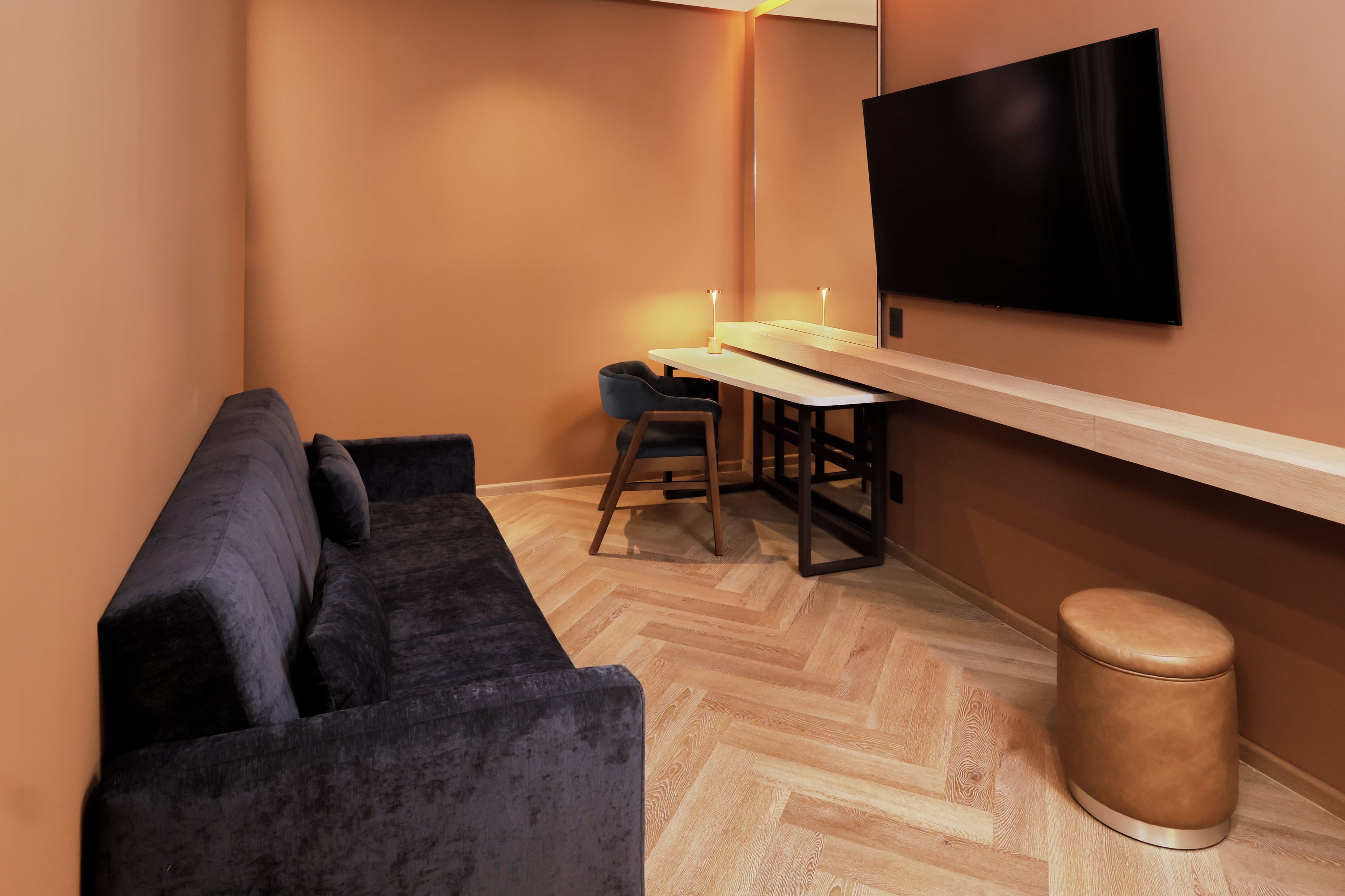 A compact living space with a sofa, a small table, a chair, a stool, and a flat-screen TV mounted on the wall.