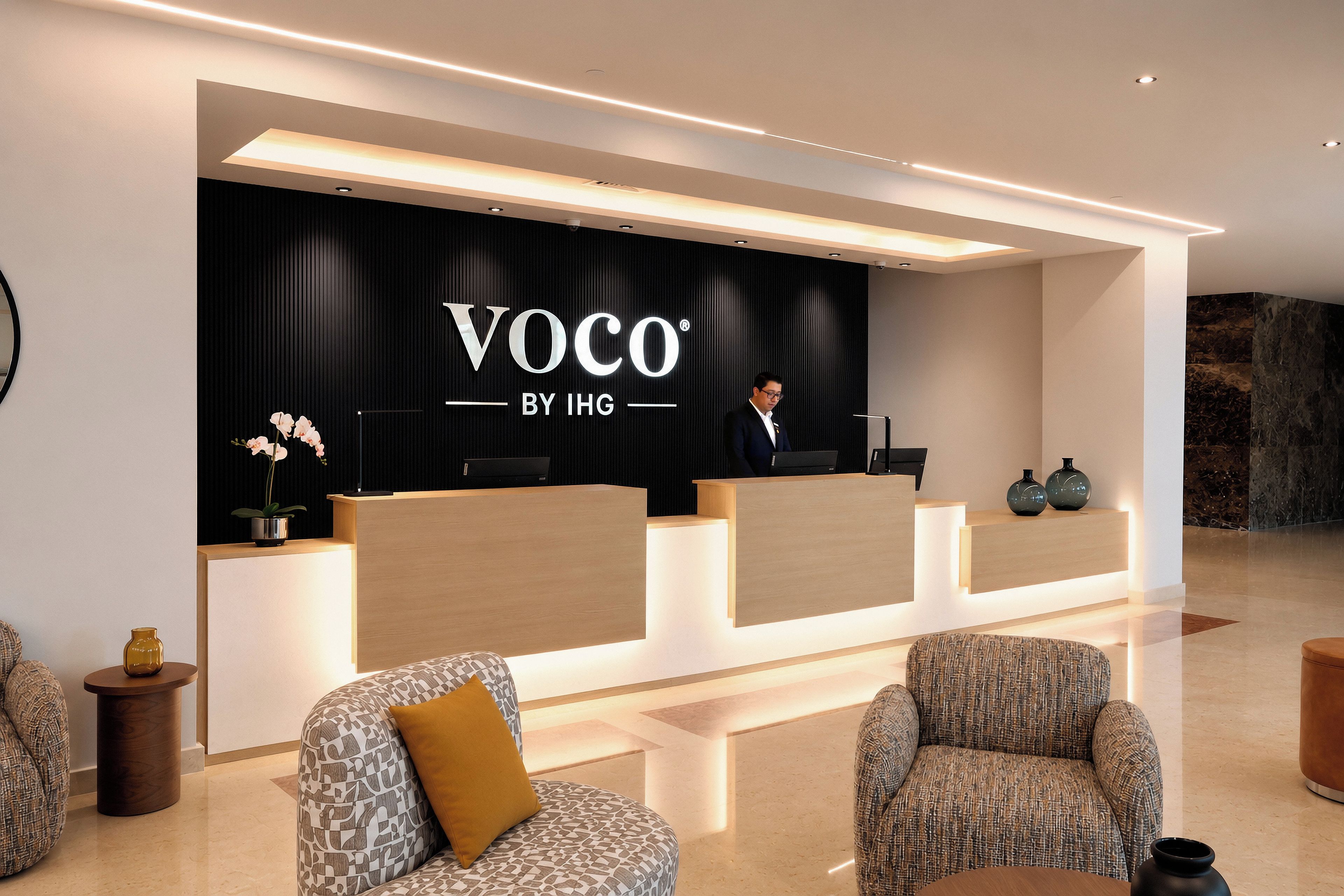A reception area with a black backdrop displaying 'Voco by IHG' branding, a receptionist, and two patterned armchairs.