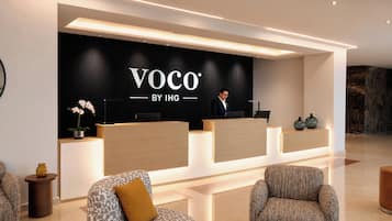 A reception area with a black backdrop displaying 'Voco by IHG' branding, a receptionist, and two patterned armchairs.