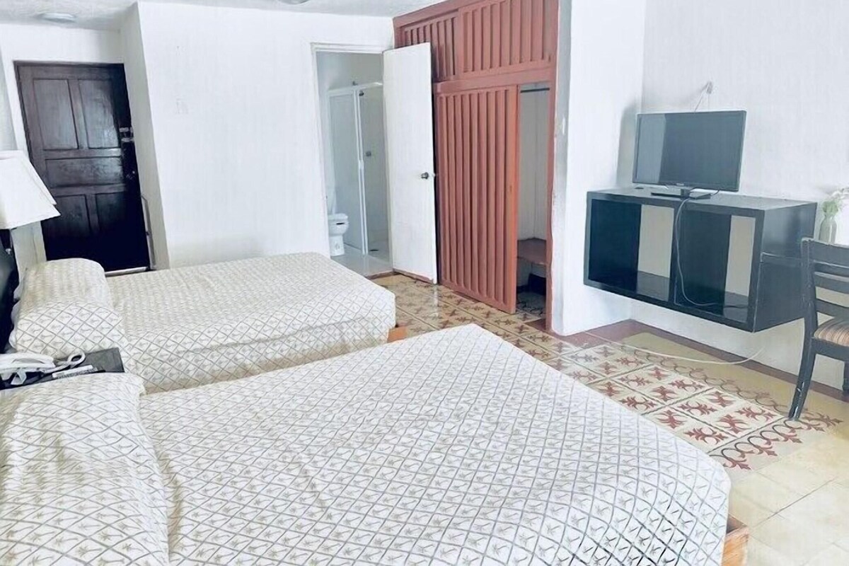 Habitación de hotel con dos camas, un escritorio con televisor, armario y ventilador de techo.