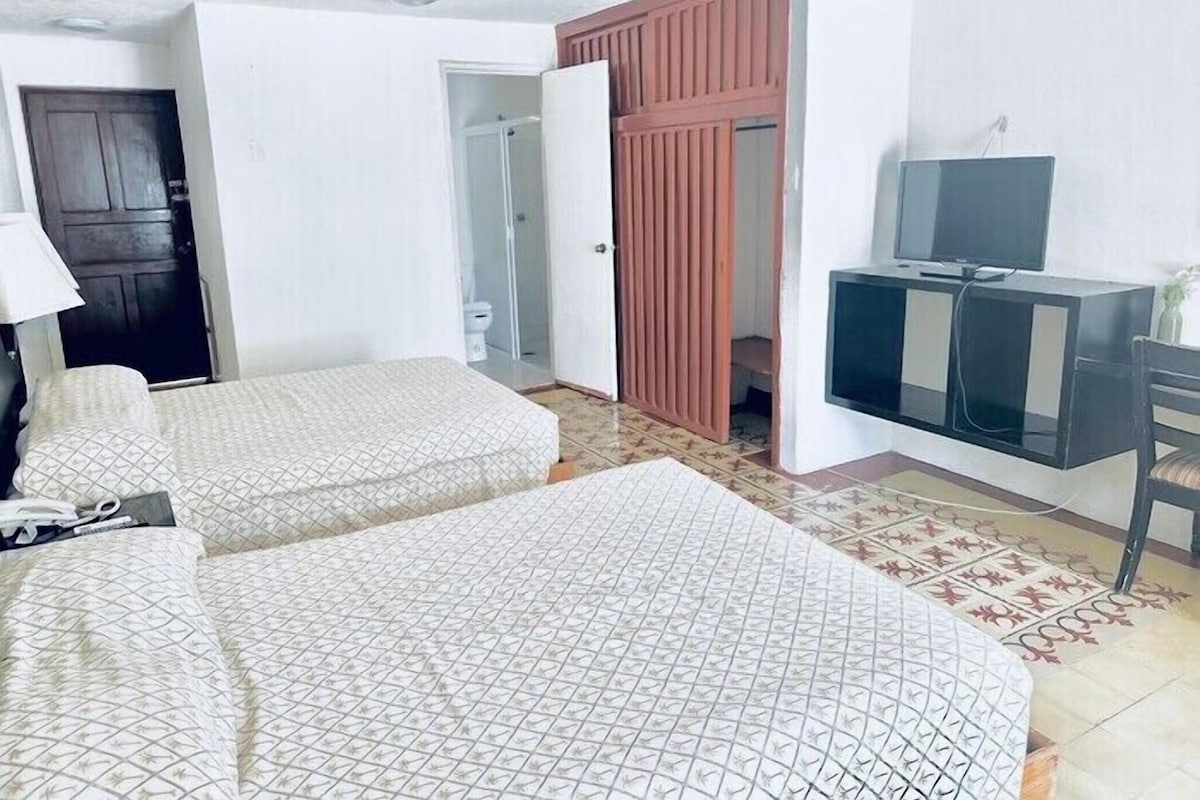 A hotel room with two beds, a desk with a TV, a wardrobe, and a ceiling fan.