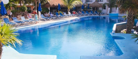 Zona de piscina con sillones de descanso y sombrillas, rodeada de palmeras y un edificio con balcones.