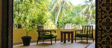 A balcony with two chairs and a small table, surrounded by a yellow wall and a view of lush greenery.