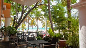An outdoor seating area with tables and chairs, surrounded by potted plants and palm trees, overlooking a beach.