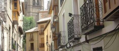 Rua estreita com edifícios de ambos os lados, uma torre de igreja destacada ao fundo e pessoas caminhando.