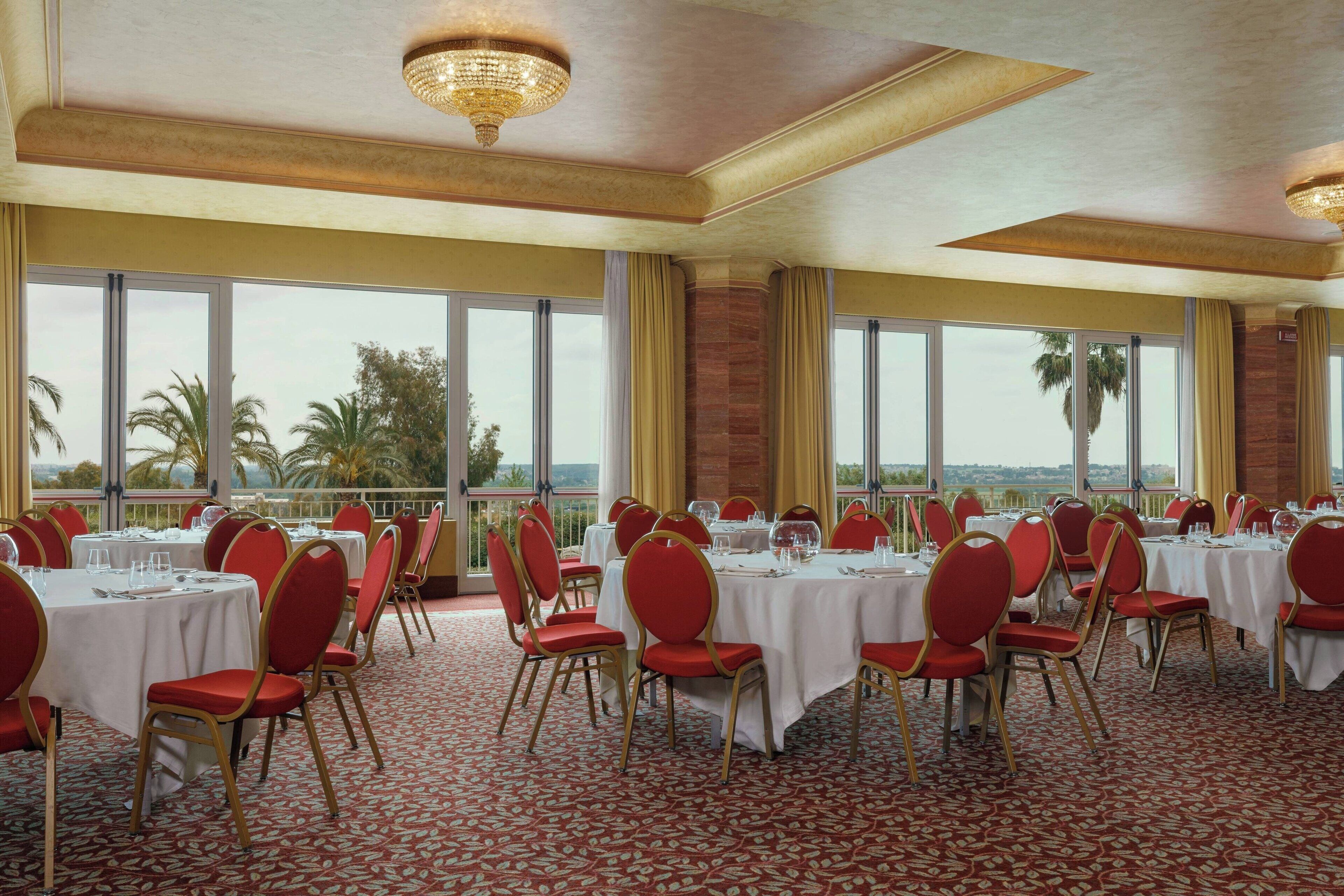 Une salle de banquet avec des tables rondes, des chaises rouges et de grandes fenêtres donnant sur des palmiers.