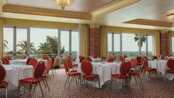 Une salle de banquet avec des tables rondes, des chaises rouges et de grandes fenêtres donnant sur des palmiers.