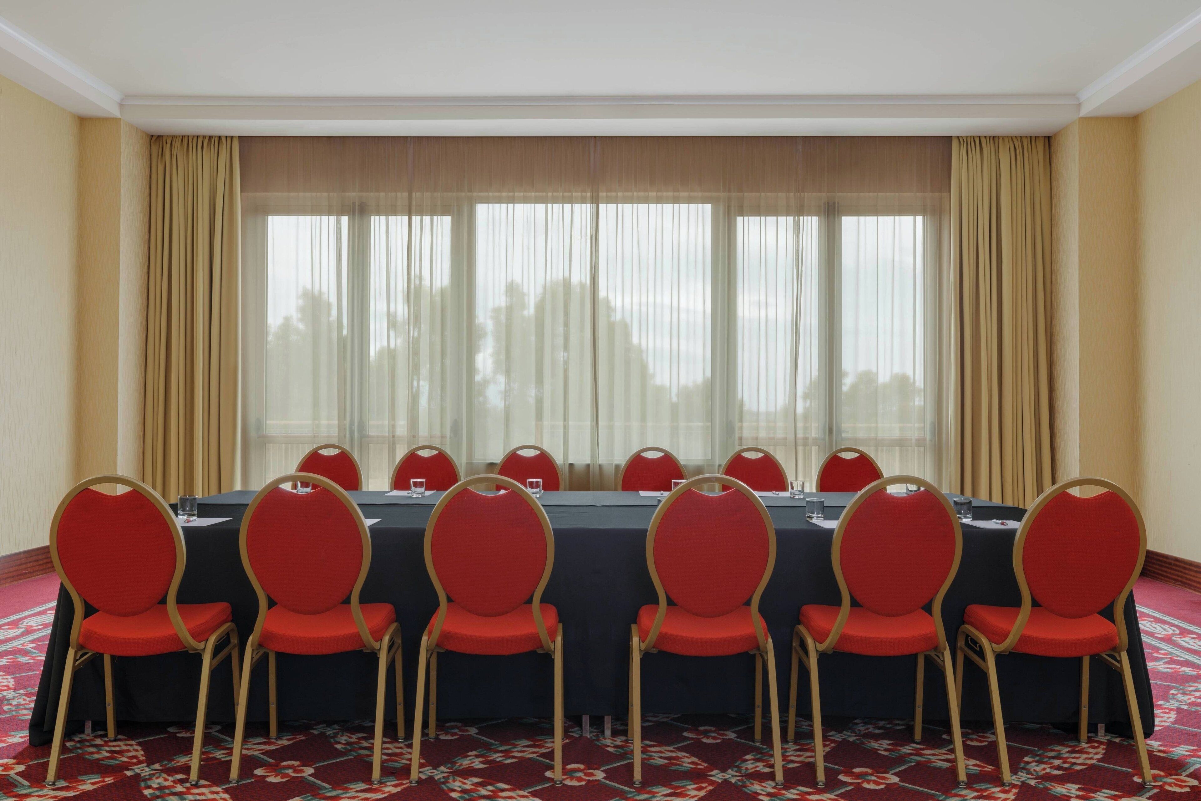 Un salón de conferencias con una larga mesa, sillas rojas y vistas amplias a través de ventanas con cortinas translúcidas.