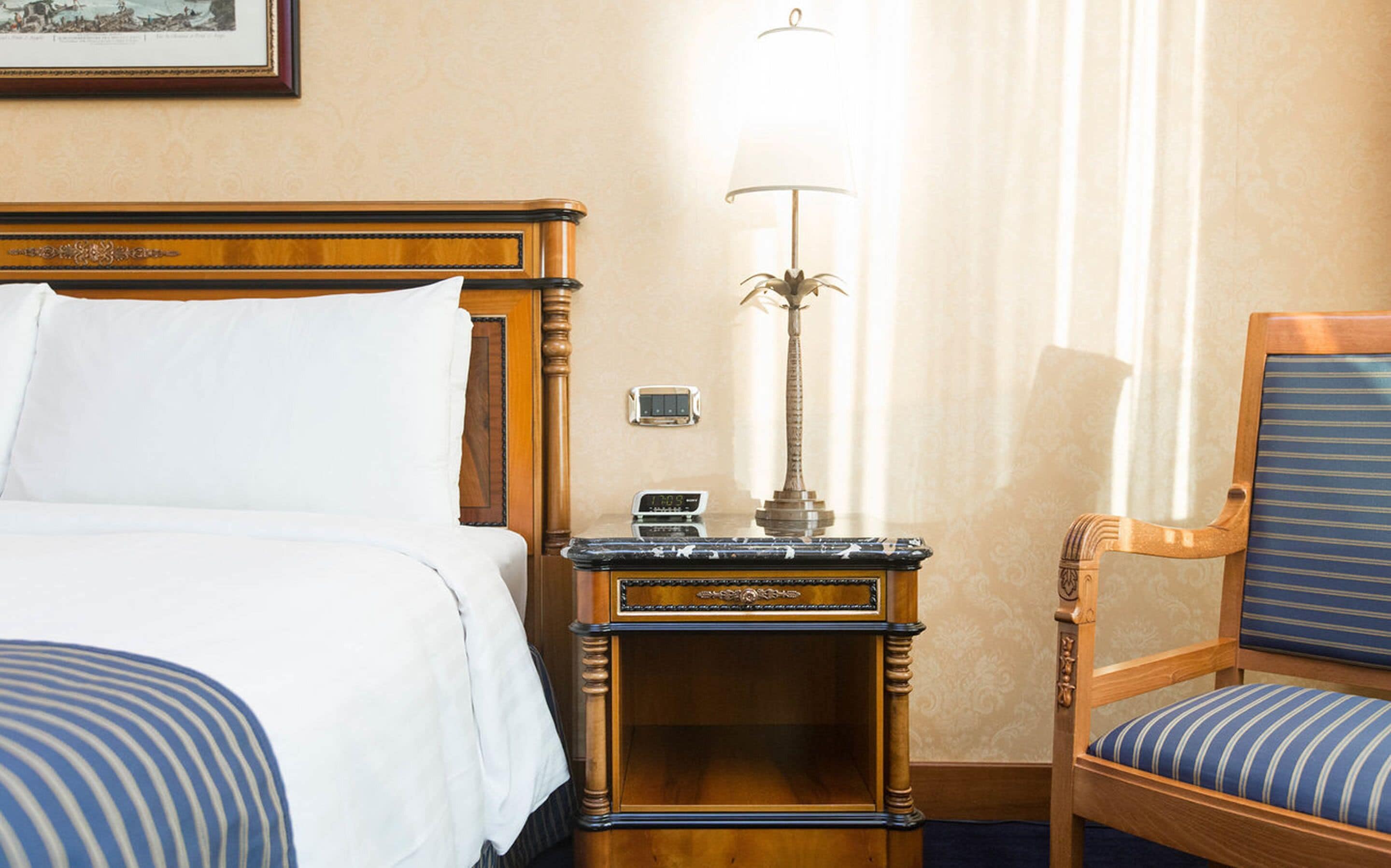 Een hotelkamer met een houten bed, een nachtkastje met een lamp, een digitale klok en een gestreepte stoel.