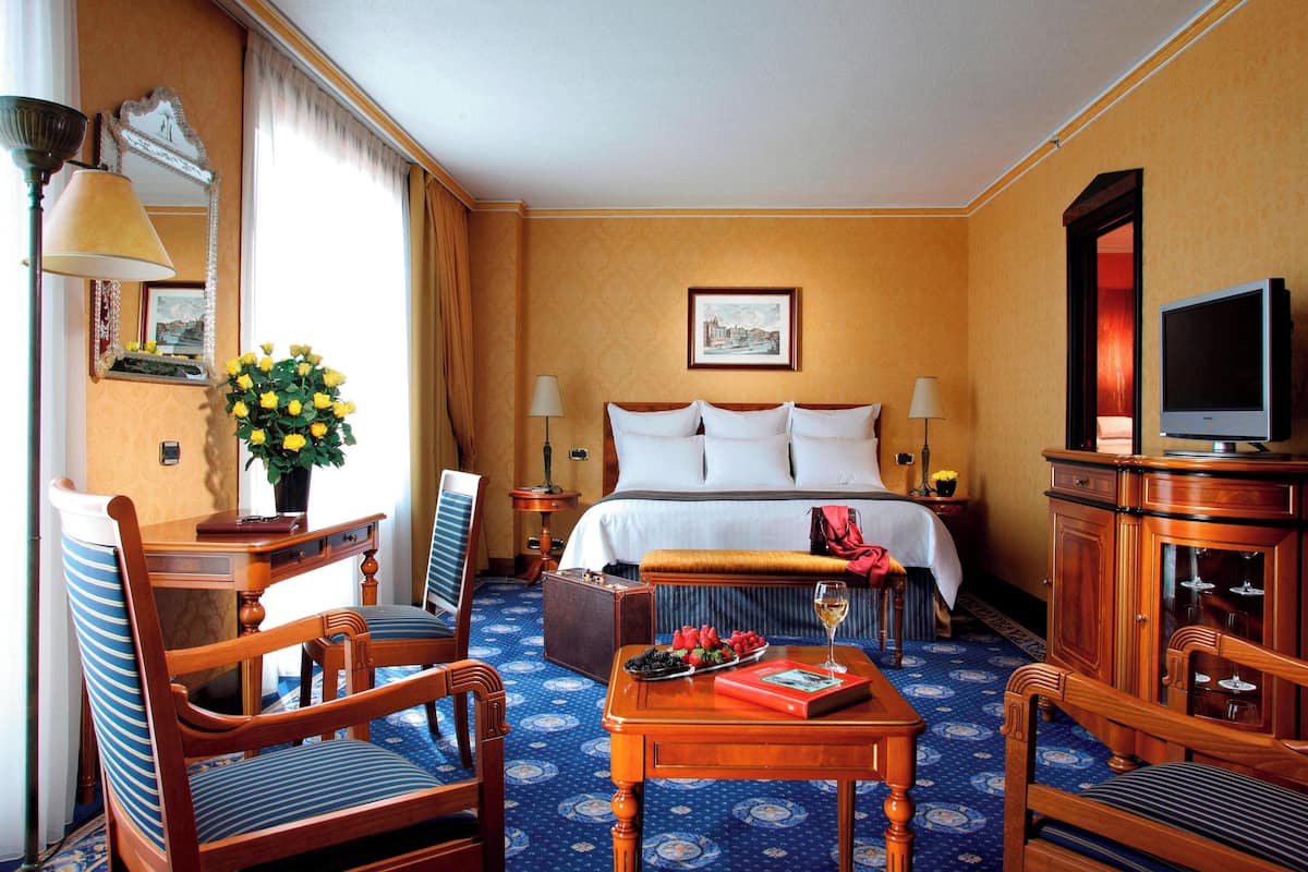 A hotel room with a bed, a desk with a lamp, a chair, a table with a book and a glass of wine, and a television.