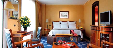 A hotel room with a bed, a desk with a lamp, a chair, a table with a book and a glass of wine, and a television.