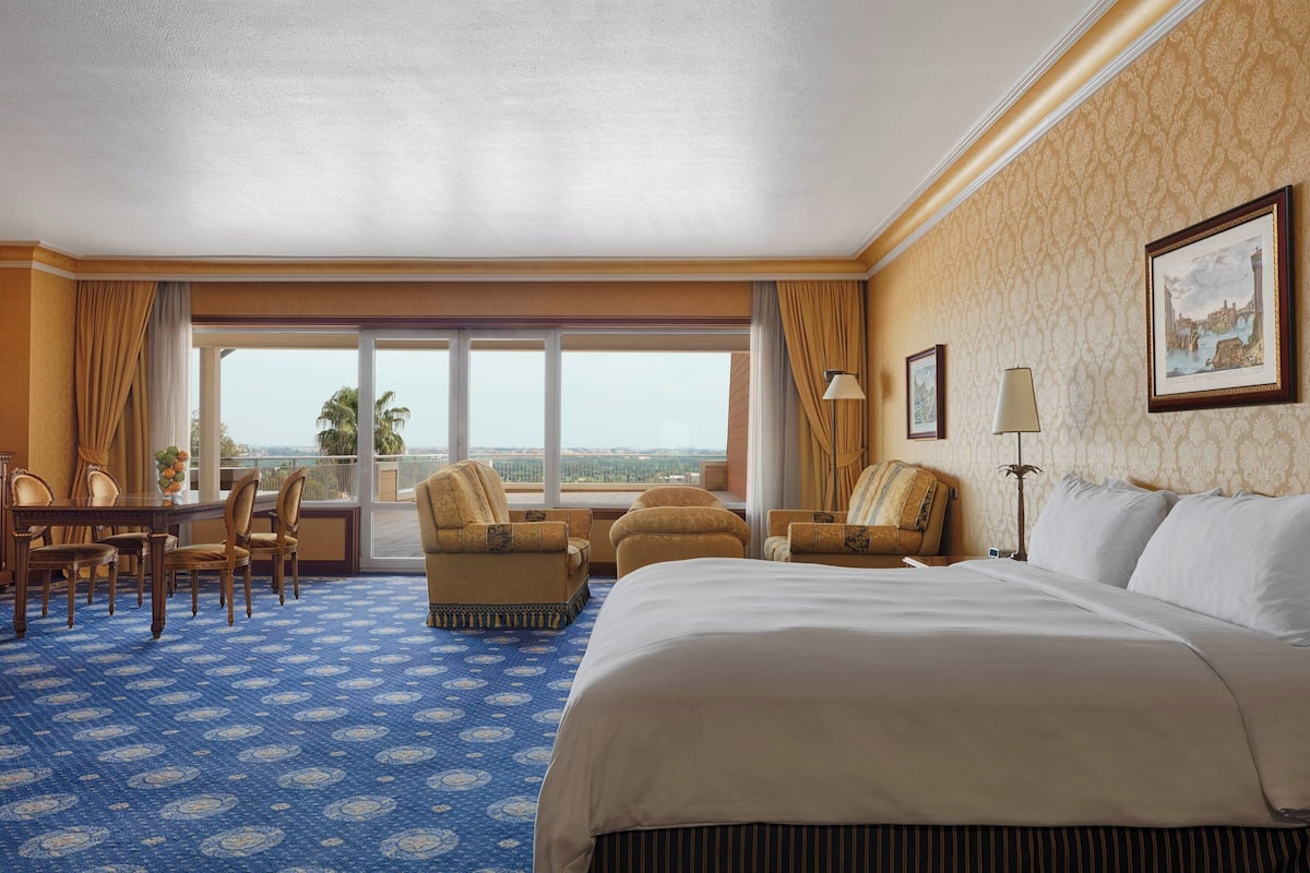 A hotel room with a large bed, a dining table with chairs, a sofa, and a view of the outdoors.