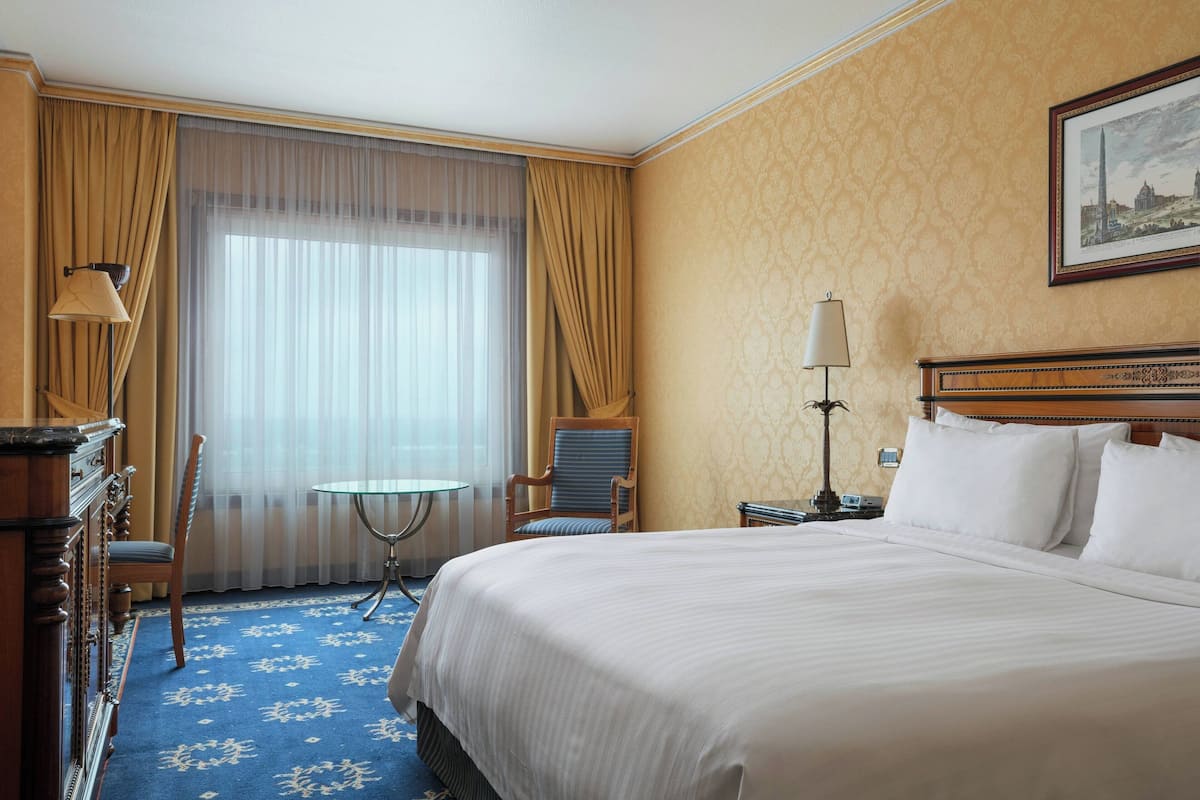 A hotel room with a large bed, a desk, a chair, a small table, and a lamp.