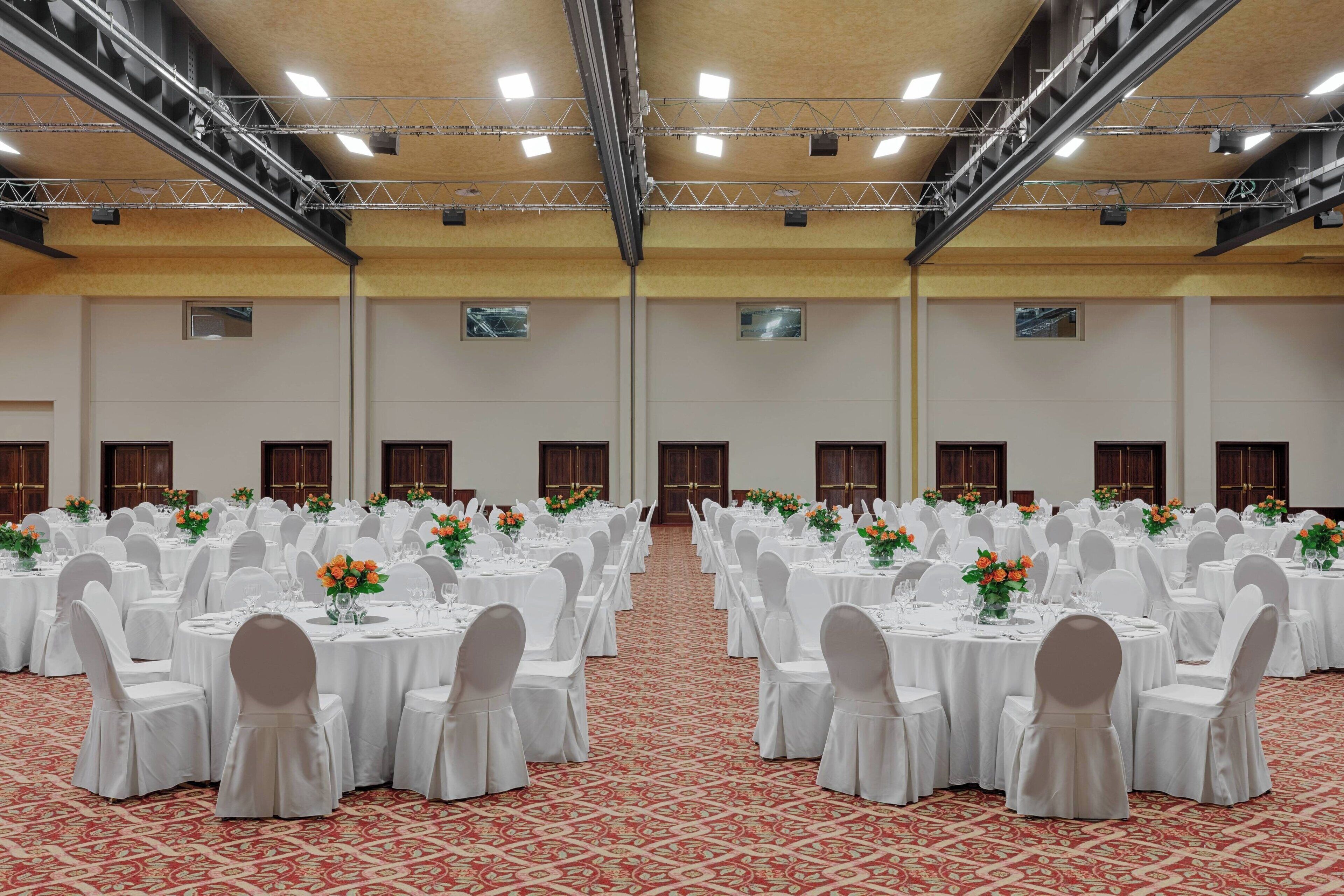 Ein großer Festsaal mit Reihen runder Tische, gedeckt mit weißen Tischdecken und Stühlen, und orangefarbenen Blumen in Vasen.