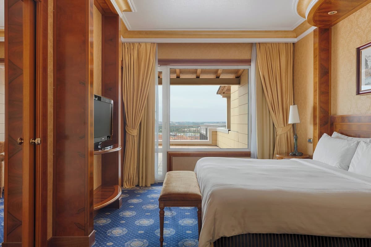 A hotel room with a large bed, a television, a bench, and a window with curtains.