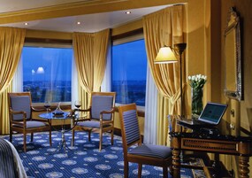 A hotel room with a large window, a desk with a laptop, two chairs, a small table with wine glasses, and a vase with flowers.