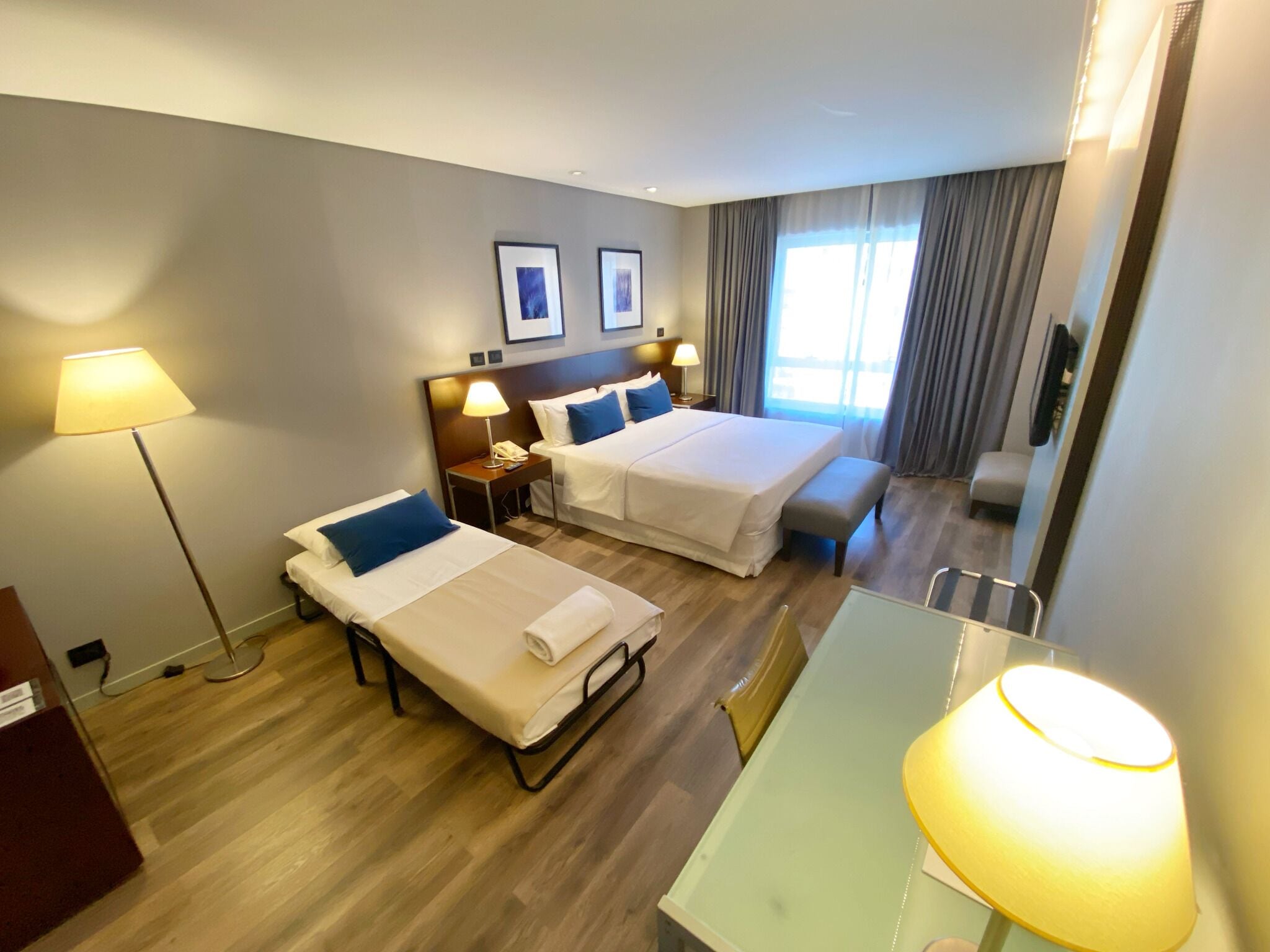 A hotel room with two single beds, a desk, and a chair. There are two bedside tables with lamps, a large mirror, and a window with curtains.