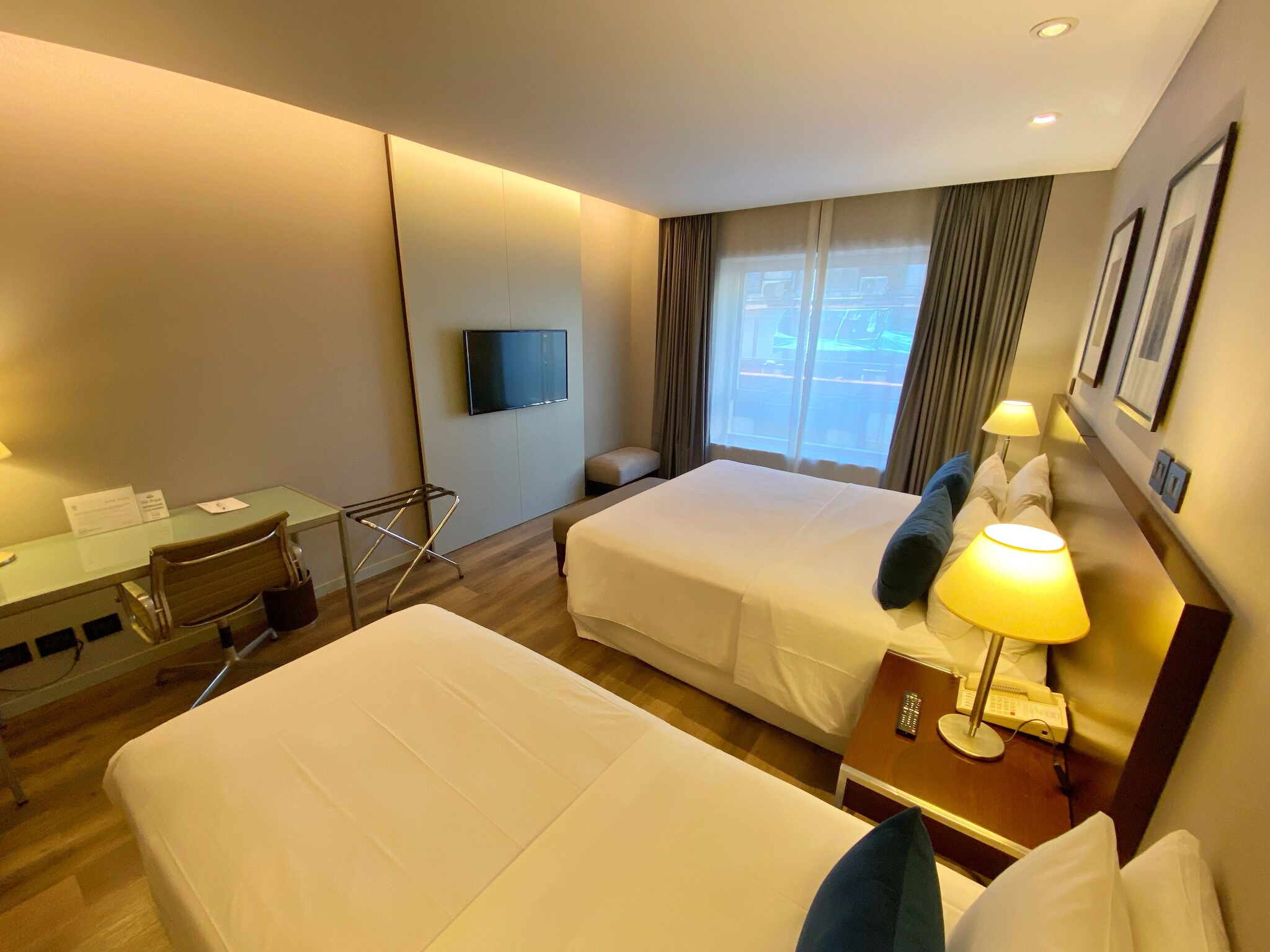 A hotel room with two beds, a desk, a chair, a TV, a window with curtains, and a bedside lamp.