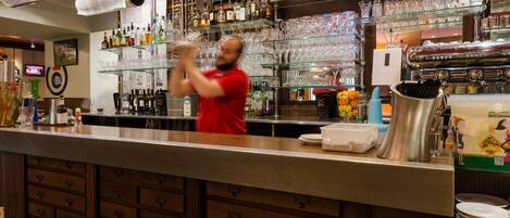 Un espace bar avec des étagères remplies de bouteilles et de verres, un comptoir où se tient une personne, et deux téléviseurs accrochés au mur.