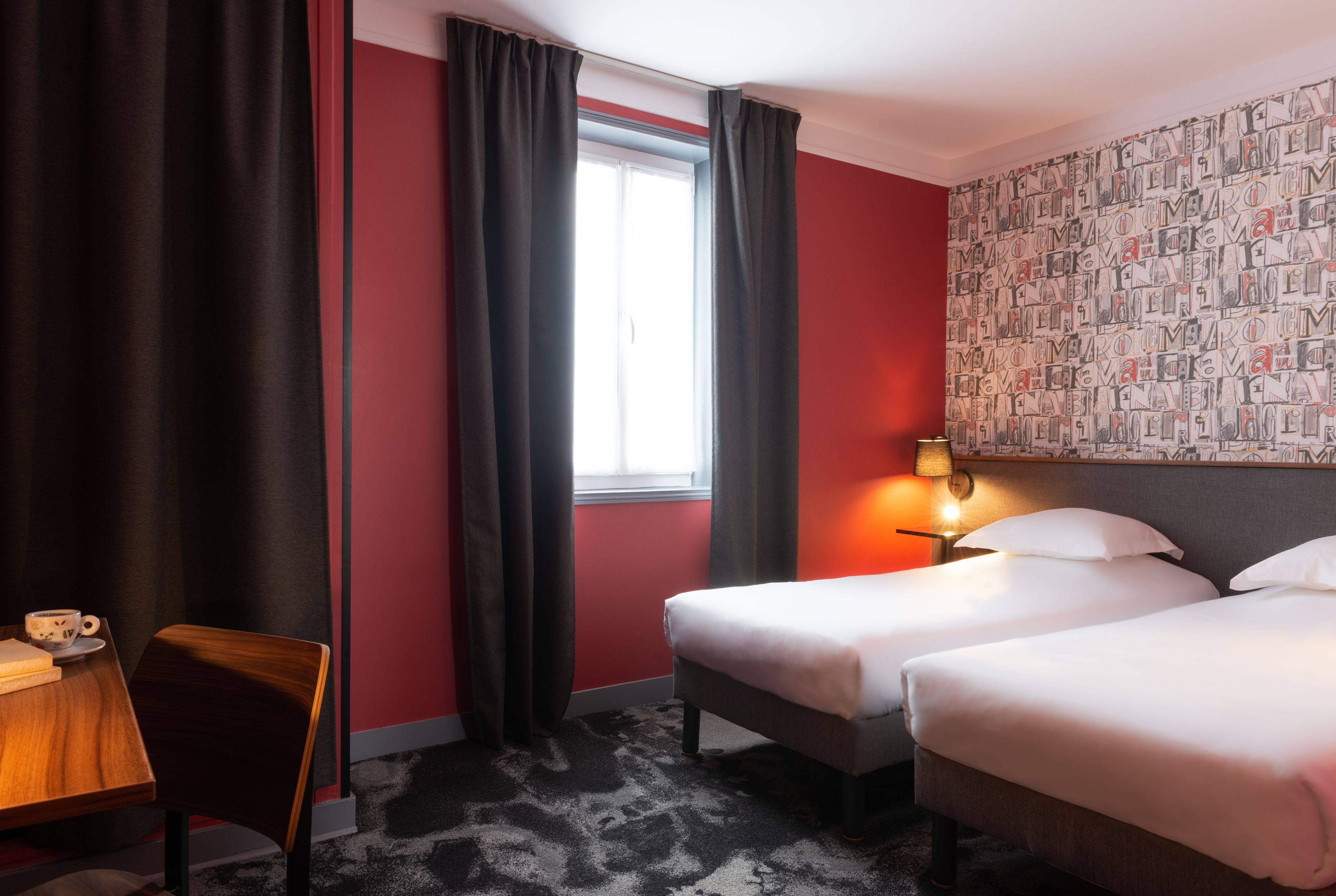 A hotel room with two beds, a desk with a chair, a window with curtains, and a wall with a patterned wallpaper.