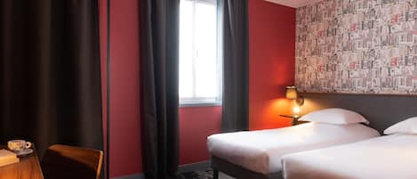 A hotel room with two beds, a desk with a chair, a window with curtains, and a wall with a patterned wallpaper.