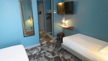 Une chambre d’hôtel avec un lit, un petit tabouret, un miroir, un téléviseur fixé au mur et une étagère avec une lampe.