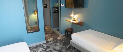 Habitación de hotel con cama, un pequeño taburete, un espejo, un televisor montado en la pared y una estantería con lámpara.