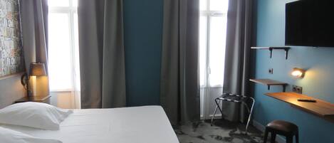 Una camera d'albergo con un letto, una scrivania, una sedia, una TV e una finestra con tende.
