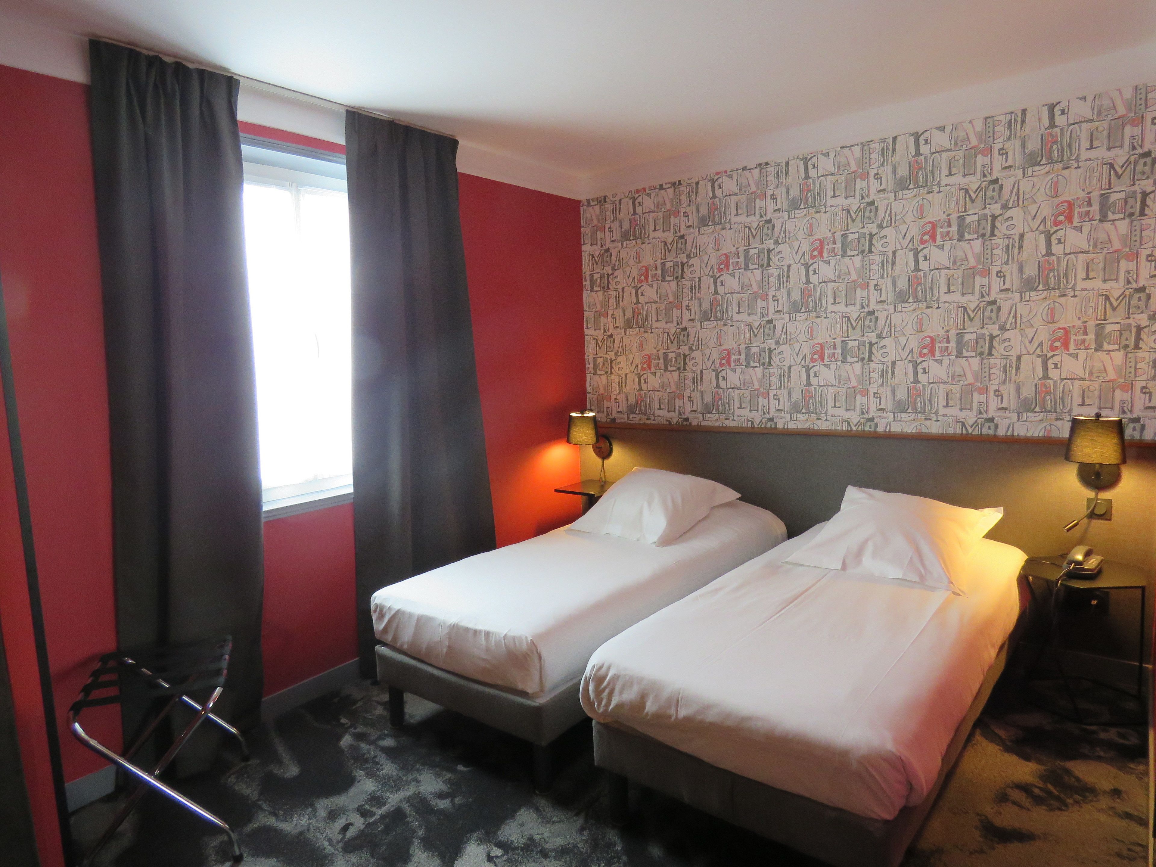 Een hotelkamer met twee enkele bedden, een rode accentmuur, behang met een patroon en een raam met gordijnen.