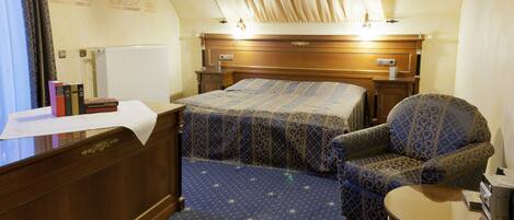 A hotel room with a large bed, a chair, a small table, and a desk with a lamp.