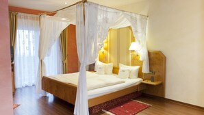 Una cama con dosel sobre cuatro postes, cabecero de madera, mesita de noche con lámpara y una alfombra roja sobre suelo de madera.