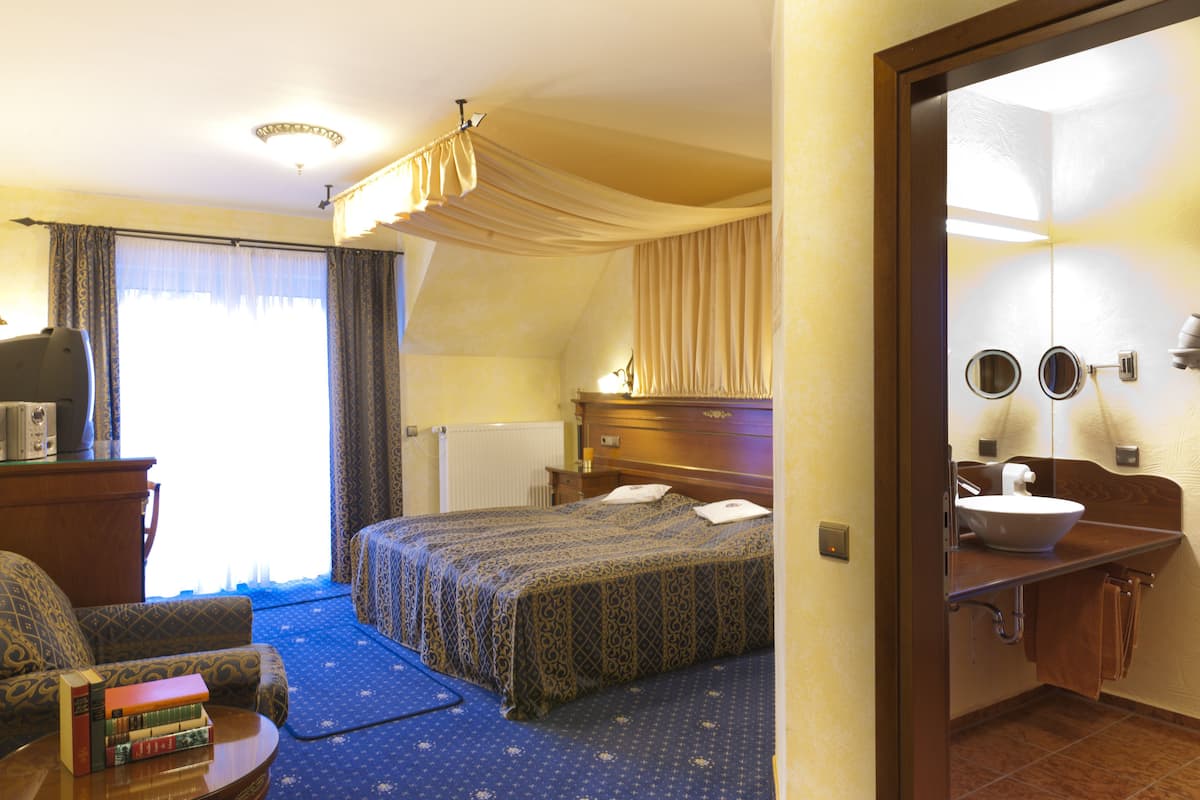 A hotel room with a bed, a desk, a chair, a bookshelf, and a bathroom with a sink and mirror.
