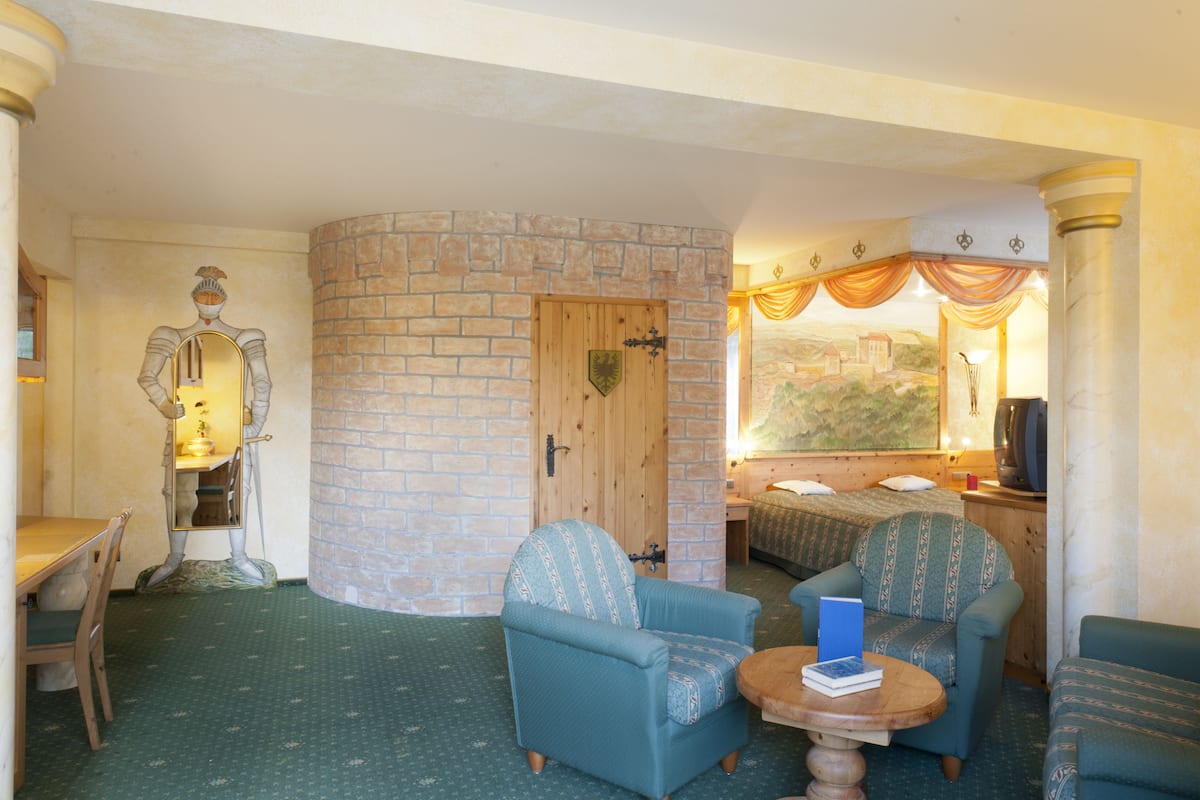 A room with a brick wall, a wooden door, a bed with a view of a landscape, a television, and a statue.
