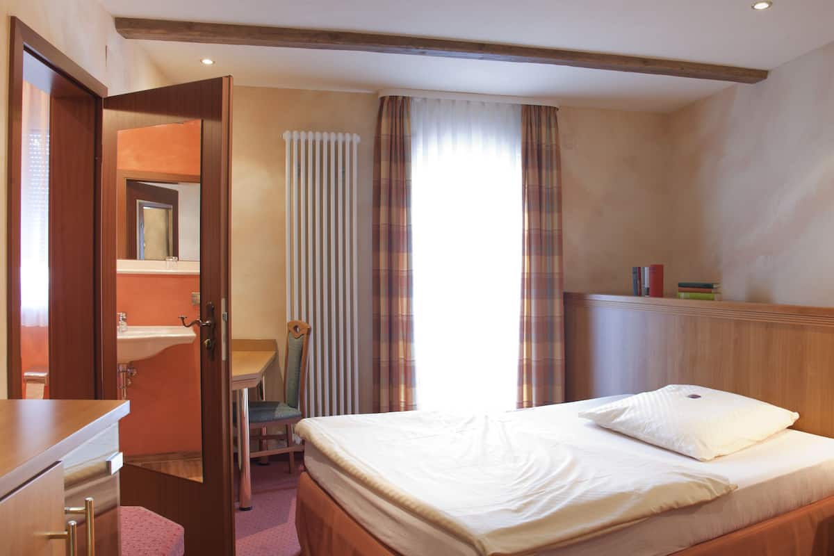 A hotel room with a bed, bedside table, a desk with a chair, a mirror, and a radiator.