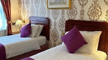 A hotel room with two beds, each with a wooden headboard, white bedding, and purple pillows. There is a framed picture on the wall and a window with curtains.