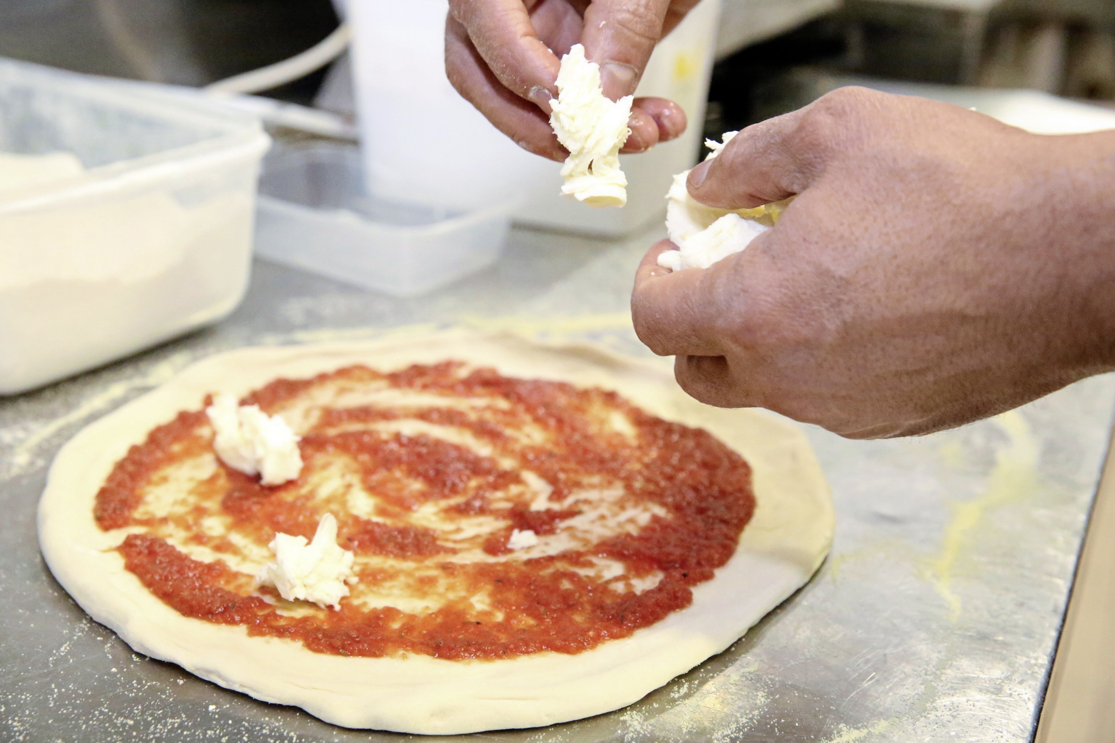 Hands preparing pizza dough with tomato sauce and cheese on a metal surface.