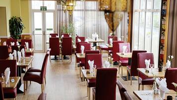 Un restaurante con sillas tapizadas en rojo y mesas de madera, dispuesto en un espacio amplio con ventanas grandes que dejan entrar luz natural.