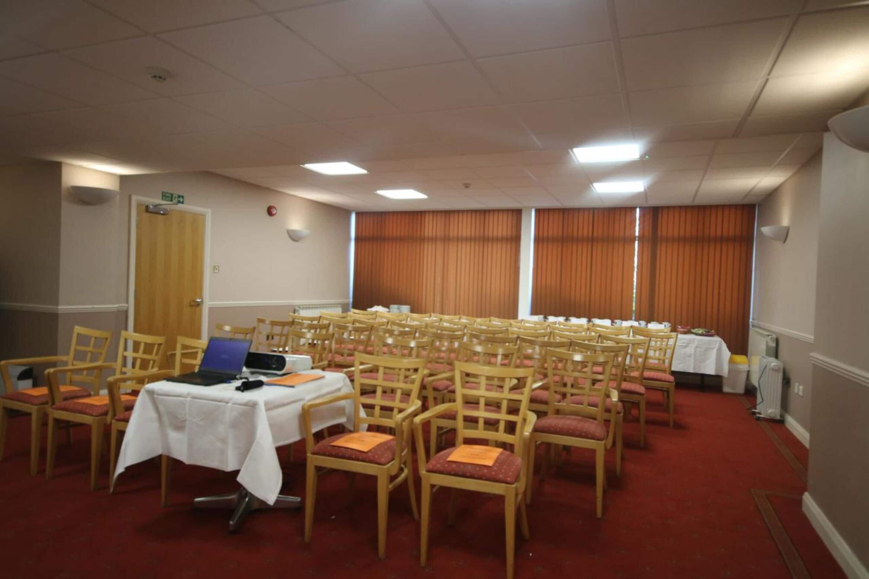 A conference room with rows of chairs, a table with a laptop, and a door with an emergency exit sign.