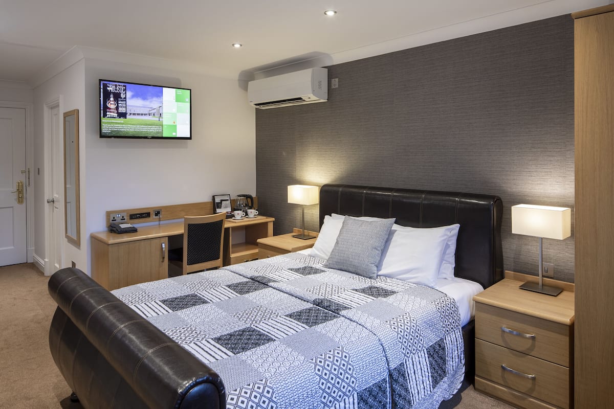 A hotel room with a bed, bedside tables, a desk, a TV, and an air conditioning unit.