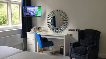 A hotel room with a bed, a desk with a computer, a blue chair, a large mirror, and a TV on the wall.