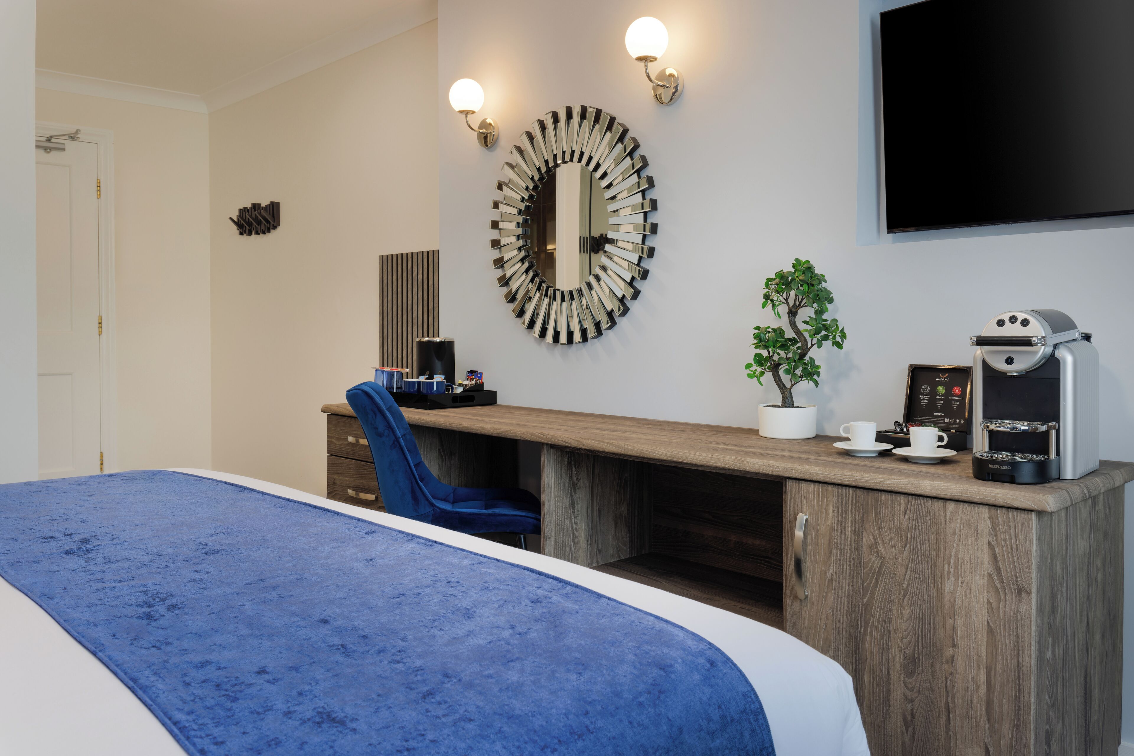 A hotel room with a large bed, a wooden desk, a blue chair, a coffee machine, a TV, and a potted plant.