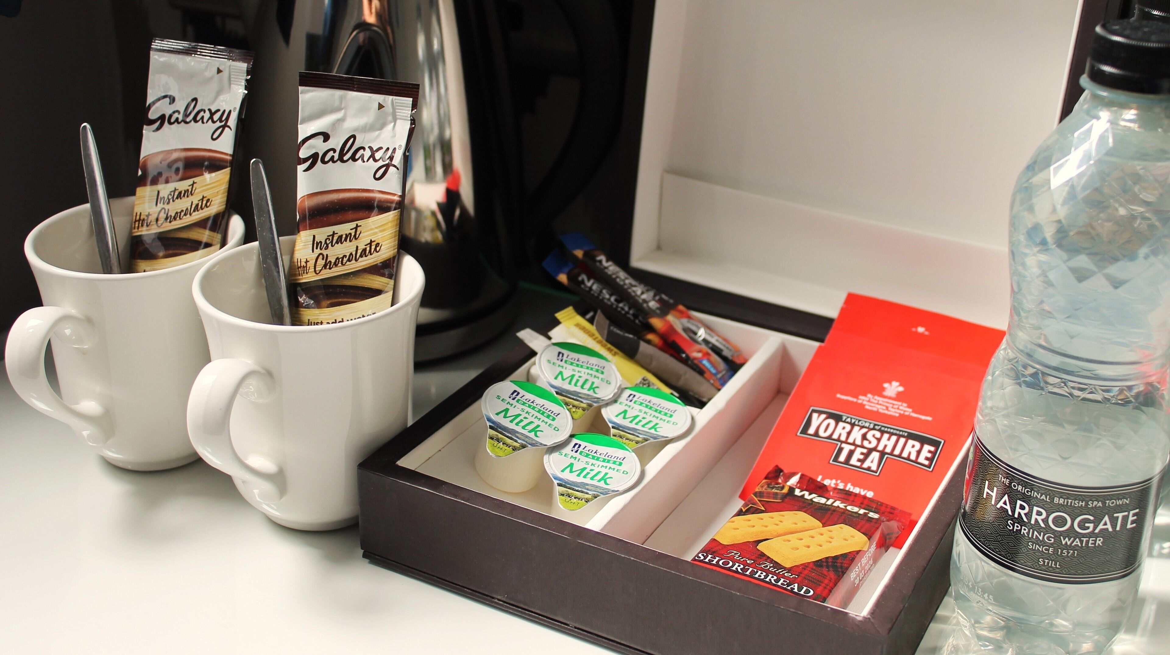 Una bandeja con sobres de chocolate caliente instantáneo, cartones de leche, té Yorkshire y una botella de agua mineral.