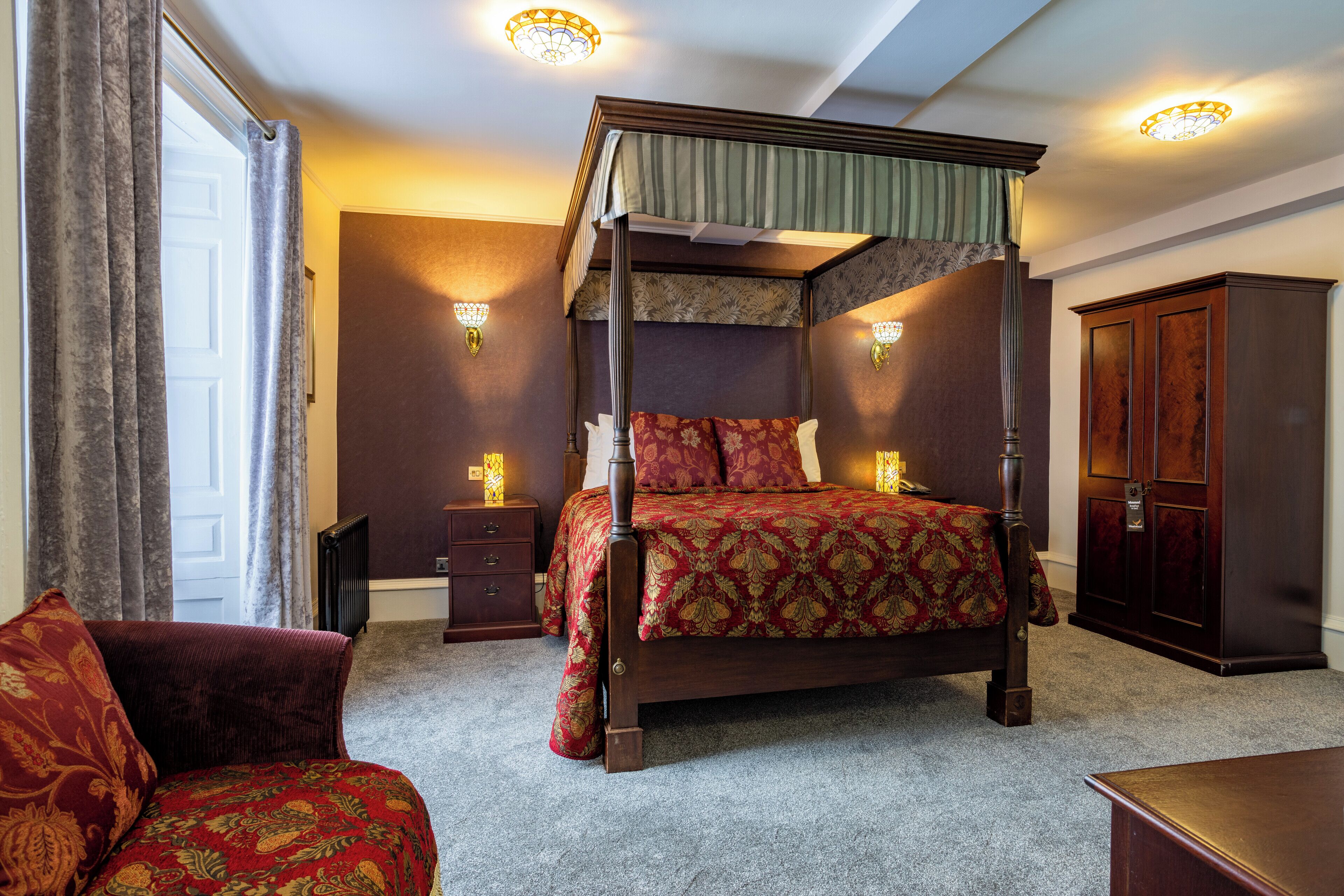 A four-poster bed with a canopy, a wooden nightstand, a dark wooden wardrobe, and a red upholstered armchair.
