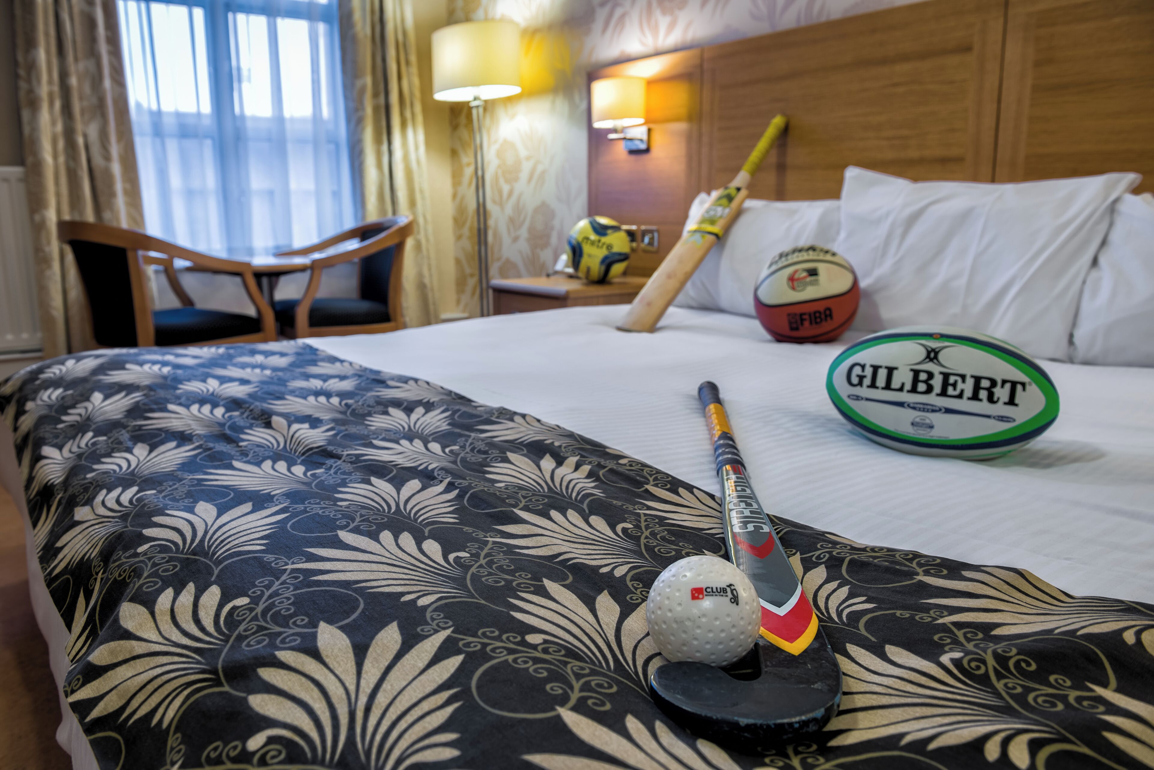 A hotel room with a bed, a desk, a chair, a lamp, and sports equipment including a cricket bat, a ball, and a rugby ball.